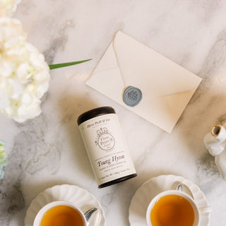 Two cups of tea with a container of tea leaves on a marble surface with flowers and a teapot.