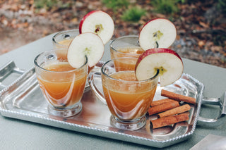 silver tray holding 4 clear glasses filled with tea, apple slices and cinnamon sticks. on a table outside